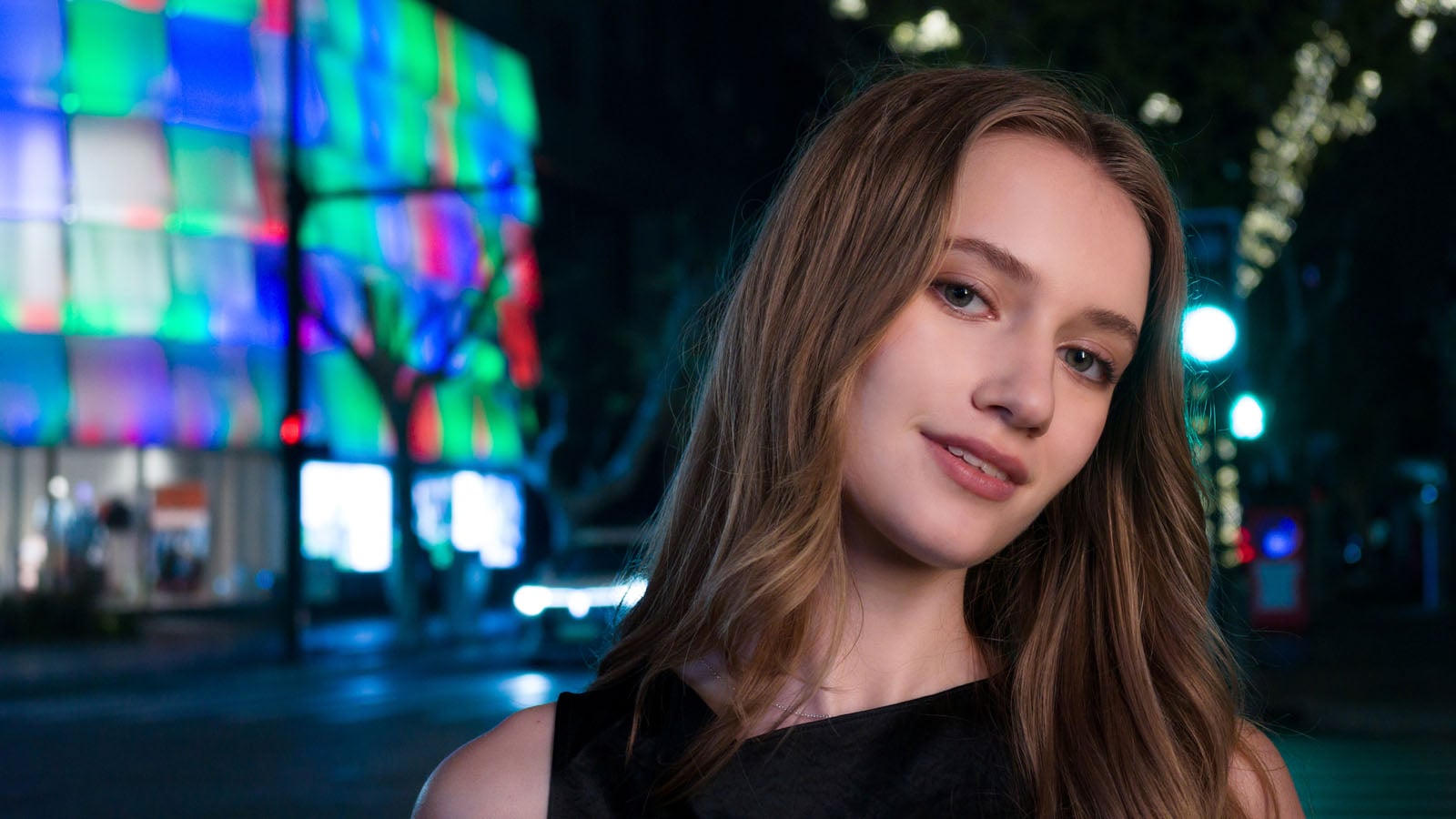 A young woman with long brown hair looks toward the camera against a blurred, colorful city street at night.