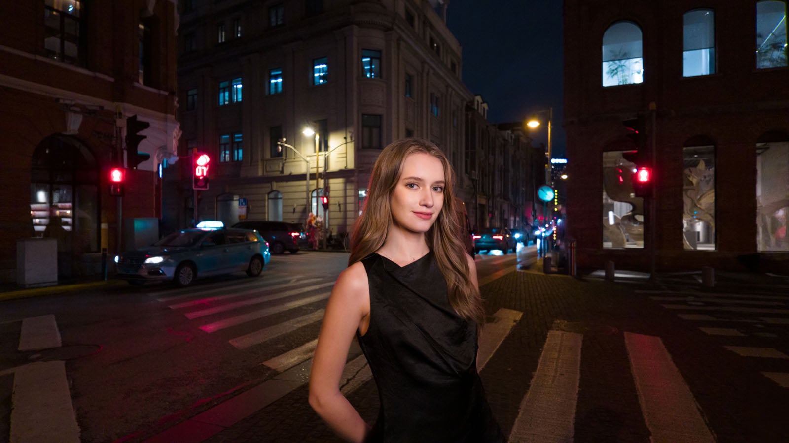 A young woman in a black dress stands on a city crosswalk at night, with streetlights, buildings, and a passing car in the background.