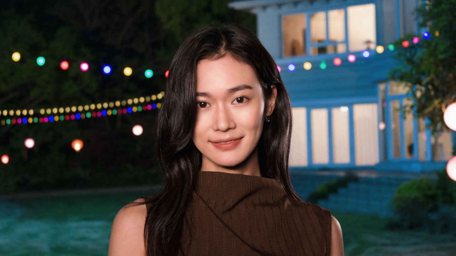 A young woman with long dark hair and a brown sleeveless top poses in front of a house decorated with colorful string lights.