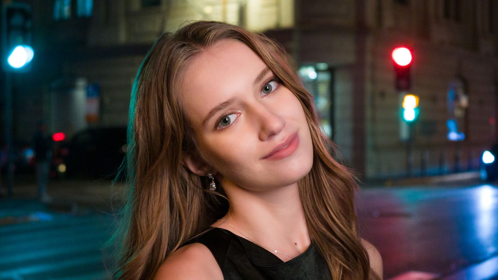 A young woman with long brown hair poses for a portrait at night against a blurred city street background with colorful traffic lights.