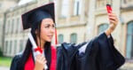 A graduate in cap and gown holds a diploma and takes a selfie outside a building, smiling proudly.
