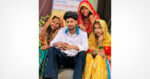A young man sits on steps, surrounded by three smiling women in colorful traditional attire and veils, posing together outdoors.