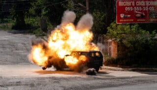 A black SUV is engulfed in a fiery explosion on an empty street, with flames and smoke billowing around the vehicle. Power lines and a red billboard are visible in the background.