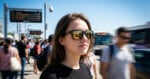 A young woman wearing sunglasses stands outdoors at a busy transit stop, with blurred people and buses moving around her on a sunny day.