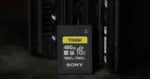 A Sony Tough CFexpress Type A memory card with 480GB capacity is standing upright in front of an electronic device, possibly a camera or card reader, on a dark surface.