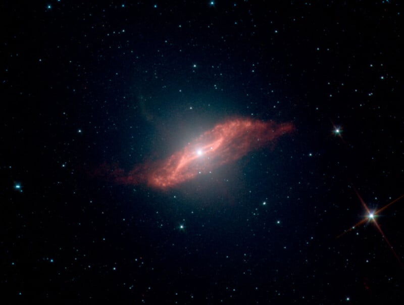 A bright spiral galaxy with a glowing, reddish-orange dust ring at its center, surrounded by numerous tiny white stars scattered across a dark space background.