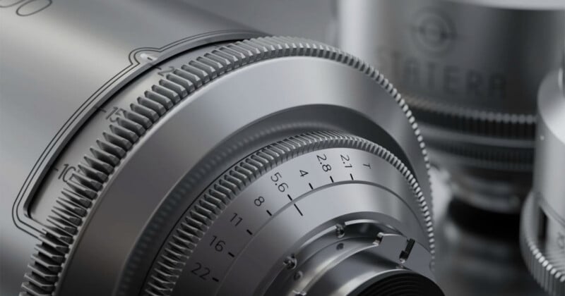Close-up of a silver camera lens with engraved numbers and textured focus rings, showing fine mechanical details and precision engineering. Other blurred lens components are visible in the background.