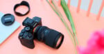 A black Sony mirrorless camera with a Tamron lens attached sits on a light pink surface next to a lens cap, a lens hood, and some green grass stalks.
