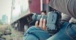 A person holding a DSLR camera sits by a train track, with a train car visible in the background. The focus is on the camera and the individual’s hand.