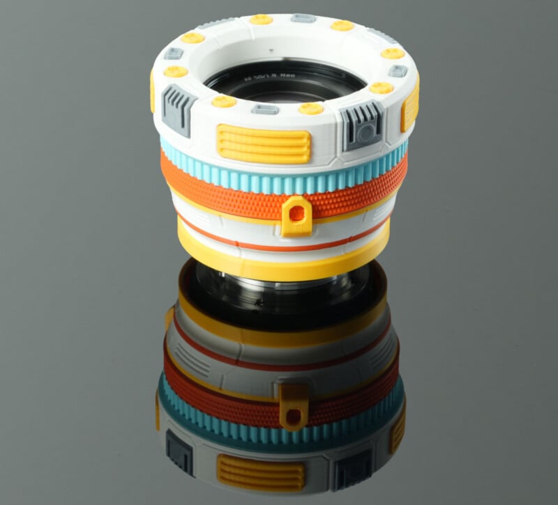 A colorful, toy-like camera lens with orange, yellow, blue, and white accents sits on a reflective black surface, showing its creative, non-traditional design.