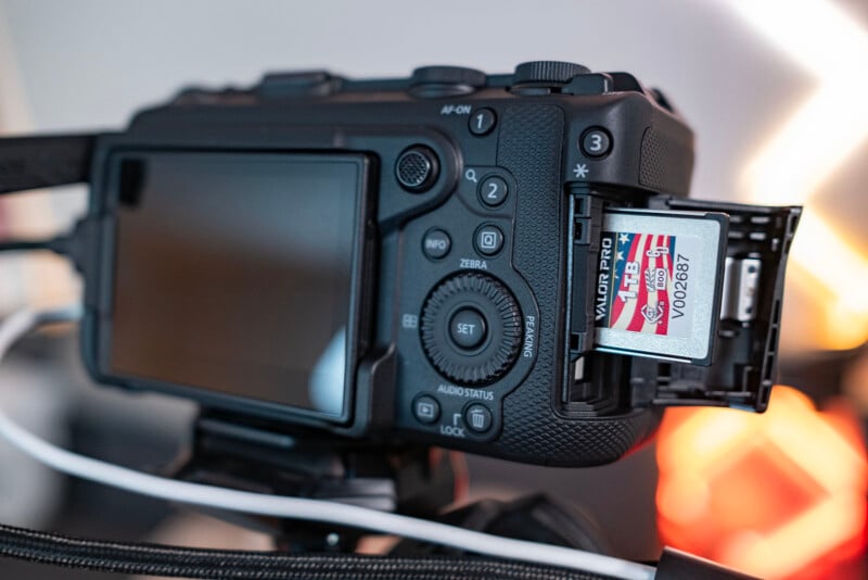 A close-up of a digital camera with its memory card slot open, showing a red and white memory card partially inserted. The camera is on a tripod and various buttons and controls are visible.