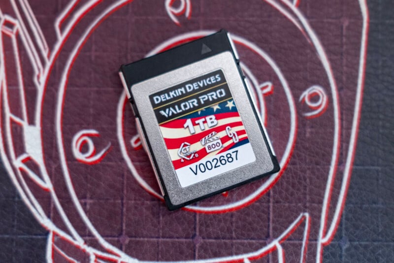 A Delkin Devices Valor Pro memory card with 1TB capacity and a label reading "V002687" is placed on a textured surface with a stylized watch design in the background.