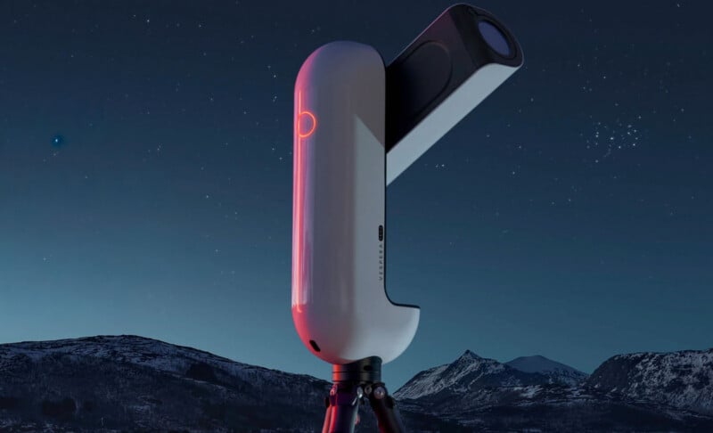 A white, modern, automated telescope stands on a tripod against a dark, starry night sky above a snowy mountain range.