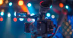 A close-up of a video camera filming in a colorful studio with bright, blurred stage lights in the background.