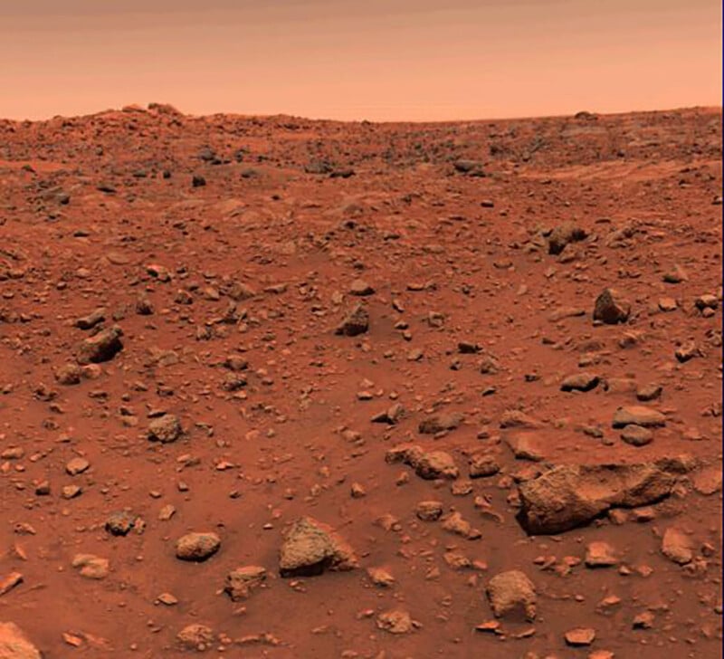 A rocky, reddish landscape with scattered stones and a dusty surface under a hazy orange sky, resembling the surface of Mars.