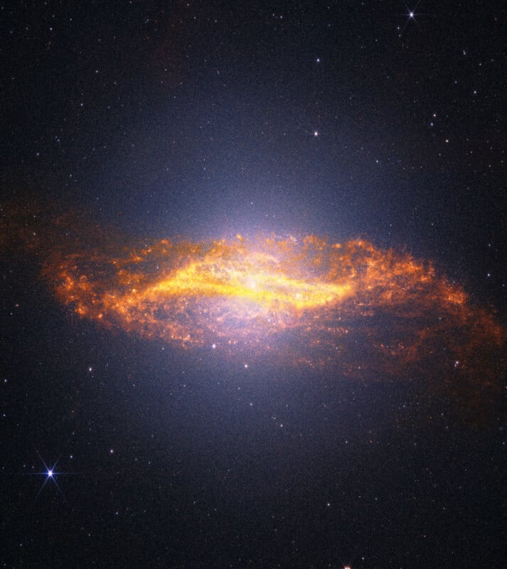 A bright, spiral galaxy glows with golden-yellow and orange light surrounded by dark space and distant stars, highlighting dust and gases within the galaxy's disk.