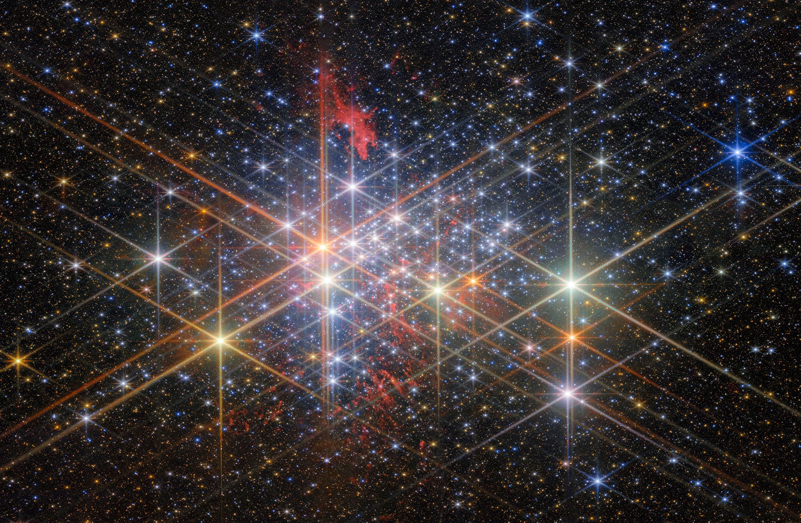 A dense star cluster sparkles with bright, colorful stars and diffraction spikes against a dark sky, with a reddish nebula visible in the upper left.