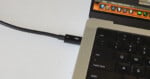 A Thunderbolt cable is plugged into a port on a laptop, next to the keyboard and screen showing application icons. The setup is on a white surface.