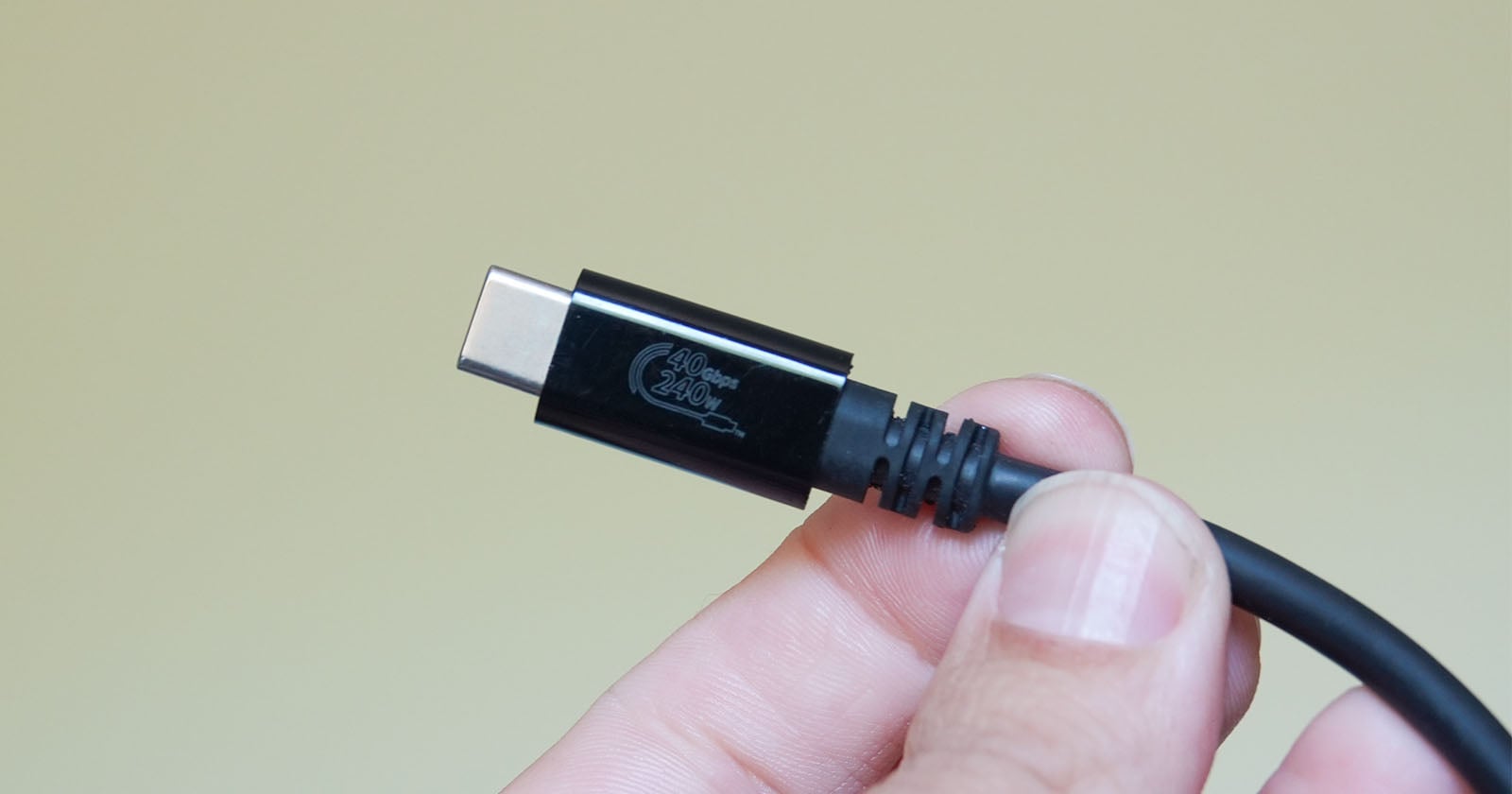 A hand holding a black USB-C cable against a plain light yellow background. The cable has a "100W 20V" marking on its connector.
