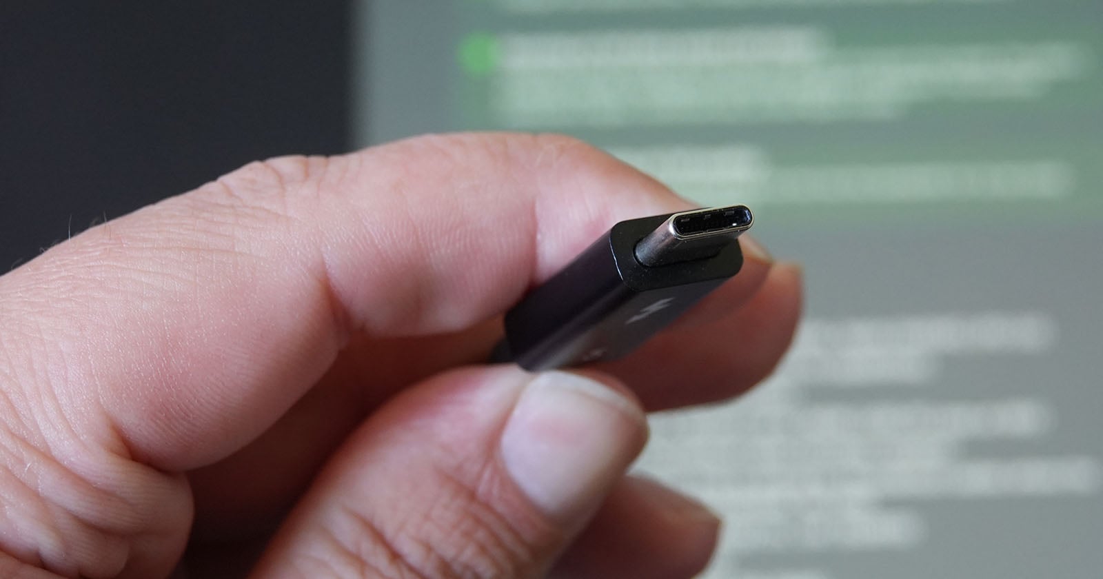 A close-up of a hand holding a USB Type-C connector, with a blurred computer screen displaying green and white text in the background.