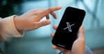 A person holds a smartphone displaying the X logo on the screen, with one hand poised to tap the device.