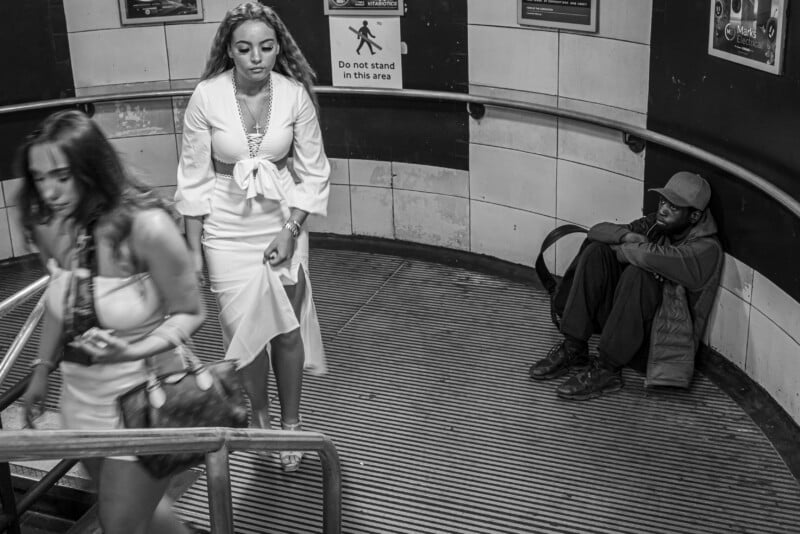 Two women in white dresses walk down subway stairs, while a man sits on the floor by the wall with his knees up, his arms resting on them. A sign reads "Do not stand in this area." The image is black and white.