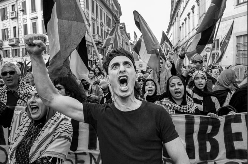 A man shouts with his fist raised during a street protest, surrounded by people holding flags and banners in a city. The image is in black and white, capturing intense emotion and solidarity.