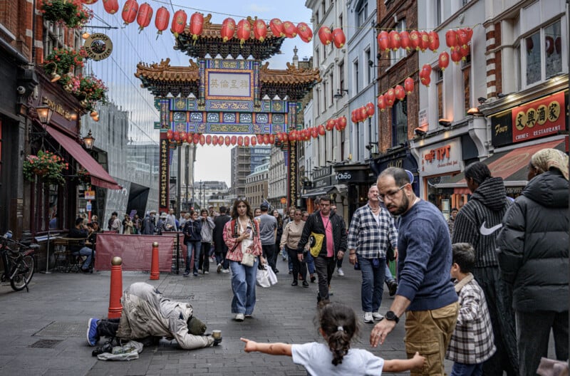 A busy Chinatown street with colorful lanterns, people walking, a child with open arms, a man reaching toward her, and a person kneeling on the ground in the foreground. Ornate gate and shops line the background.