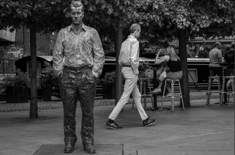 A realistic statue of a man stands with hands in pockets while people walk and sit nearby in an outdoor urban area with trees.
