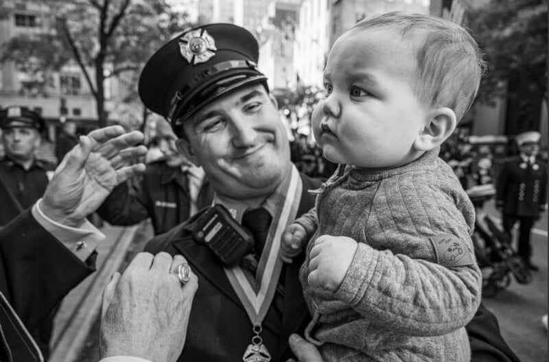 A smiling firefighter in uniform holds a baby at a parade or public event, with other uniformed people and buildings in the background. The baby looks serious and gazes off to the side. The image is in black and white.