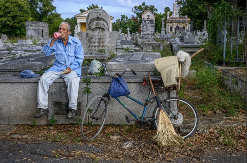 An older man in light blue clothes sits on a tomb, drinking from a pink bottle. Nearby, an old bicycle with bags, a mop, and a broom leans against the gravestone in a cemetery.