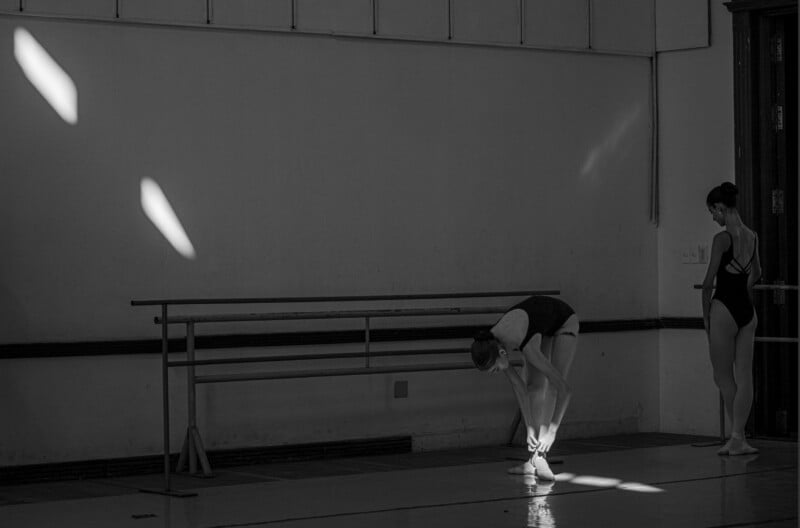 Two ballet dancers in a dim studio; one adjusts her shoe under beams of light, while the other stands nearby watching. A ballet barre and wall are in the background.