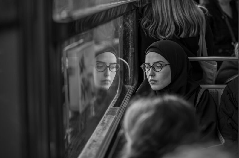 A woman in a hijab and glasses looks out a bus window, her face reflected in the glass. She appears thoughtful, surrounded by other passengers. The photo is in black and white.
