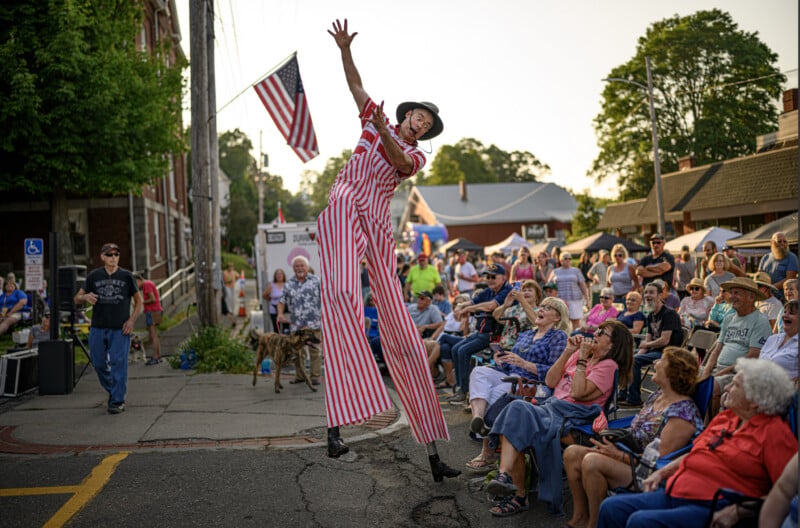 A performer on stilts in red and white stripes entertains a seated crowd at an outdoor event. An American flag is visible, and people watch, smiling, on a sunny day.