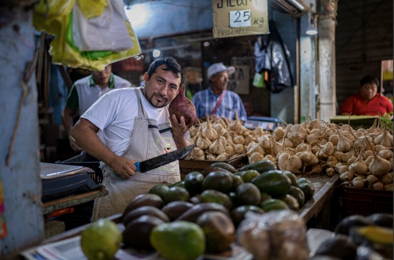 A vendor in a market smiles while holding a large root vegetable and a knife, surrounded by avocados and other produce. Signs and other people are visible in the background.