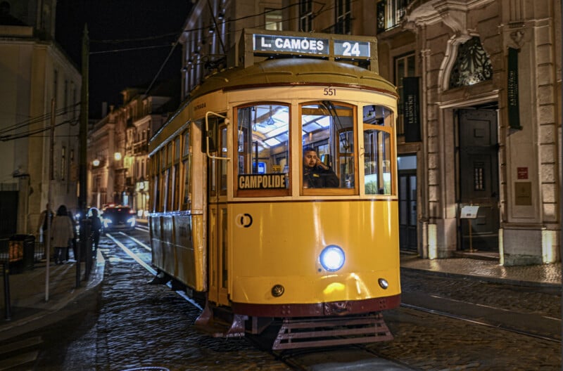 A yellow tram labeled "24 L. CAMOES" travels on cobblestone tracks at night in a city, with passengers inside and historic buildings lining the street.