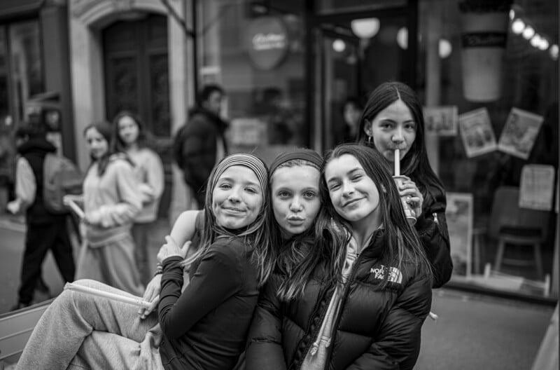 Four girls pose and smile for the camera on a city street; one girl sips a drink with a straw. Several other people are blurred in the background. The image is in black and white.