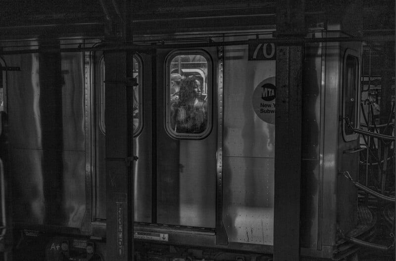 A person stands inside a dimly lit subway train, gazing out the window. The photo is in black and white, with reflections and metal bars visible on the platform outside.