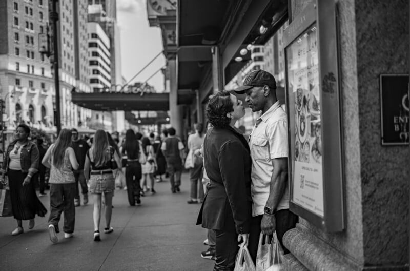 A couple stands closely facing each other on a busy city sidewalk, holding shopping bags. People walk by, and tall buildings and storefronts line the street. The image is in black and white.