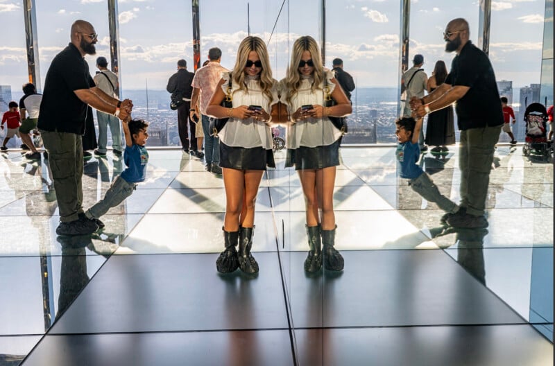 A woman with blonde hair and sunglasses looks at her phone, standing on a reflective floor with her mirror image beside her. Other people and reflections are visible in the bright, modern space.
