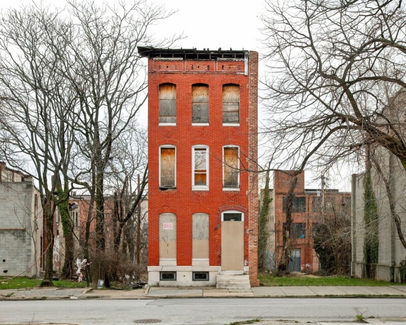 A narrow, three-story red brick building stands vacant with boarded-up windows and doors, surrounded by bare trees.