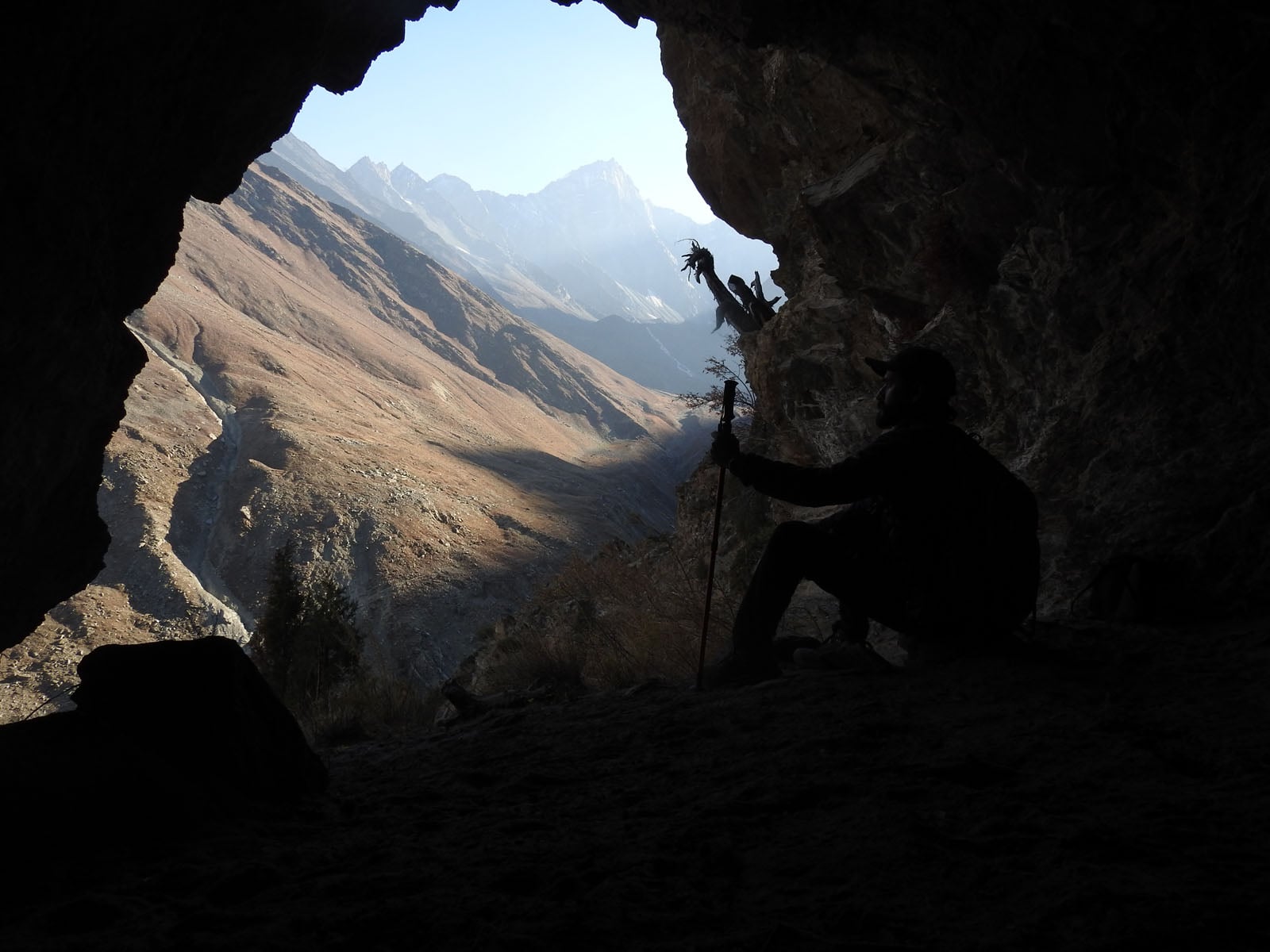 A silhouette of a person holding a hiking pole sits inside a dark cave, looking out over a sunlit mountain valley.