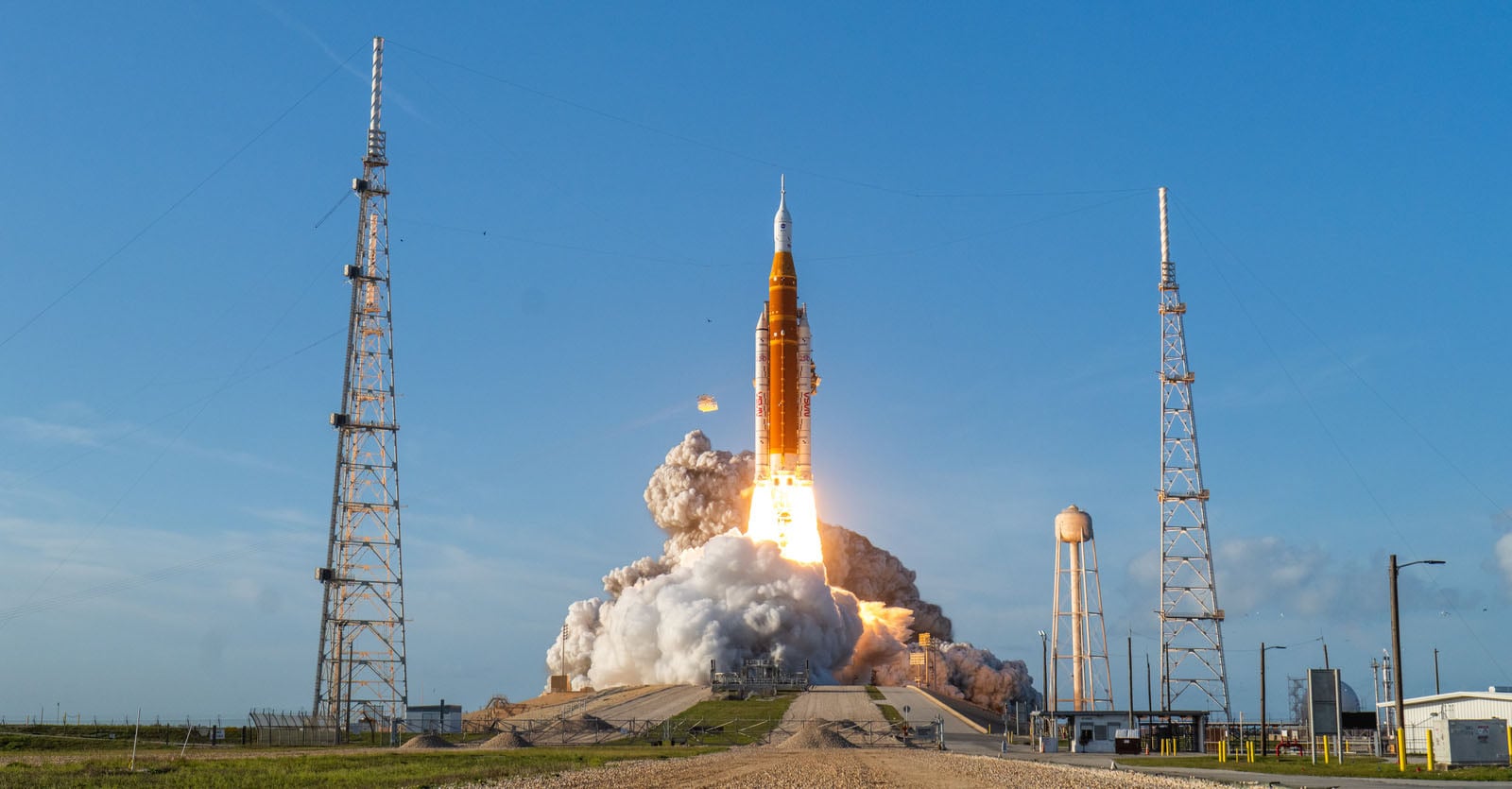 A large orange and white rocket launches into a clear blue sky, surrounded by a massive cloud of smoke and two tall towers.