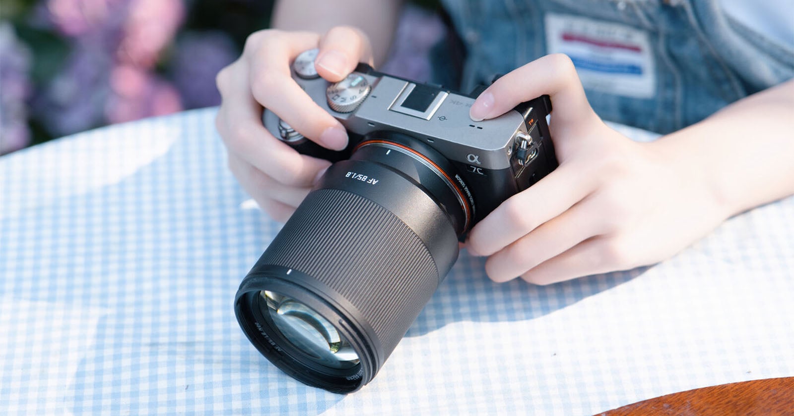 A person's hands hold a silver and black mirrorless camera with an 85mm f/1.8 lens, resting on a blue and white checkered tablecloth.
