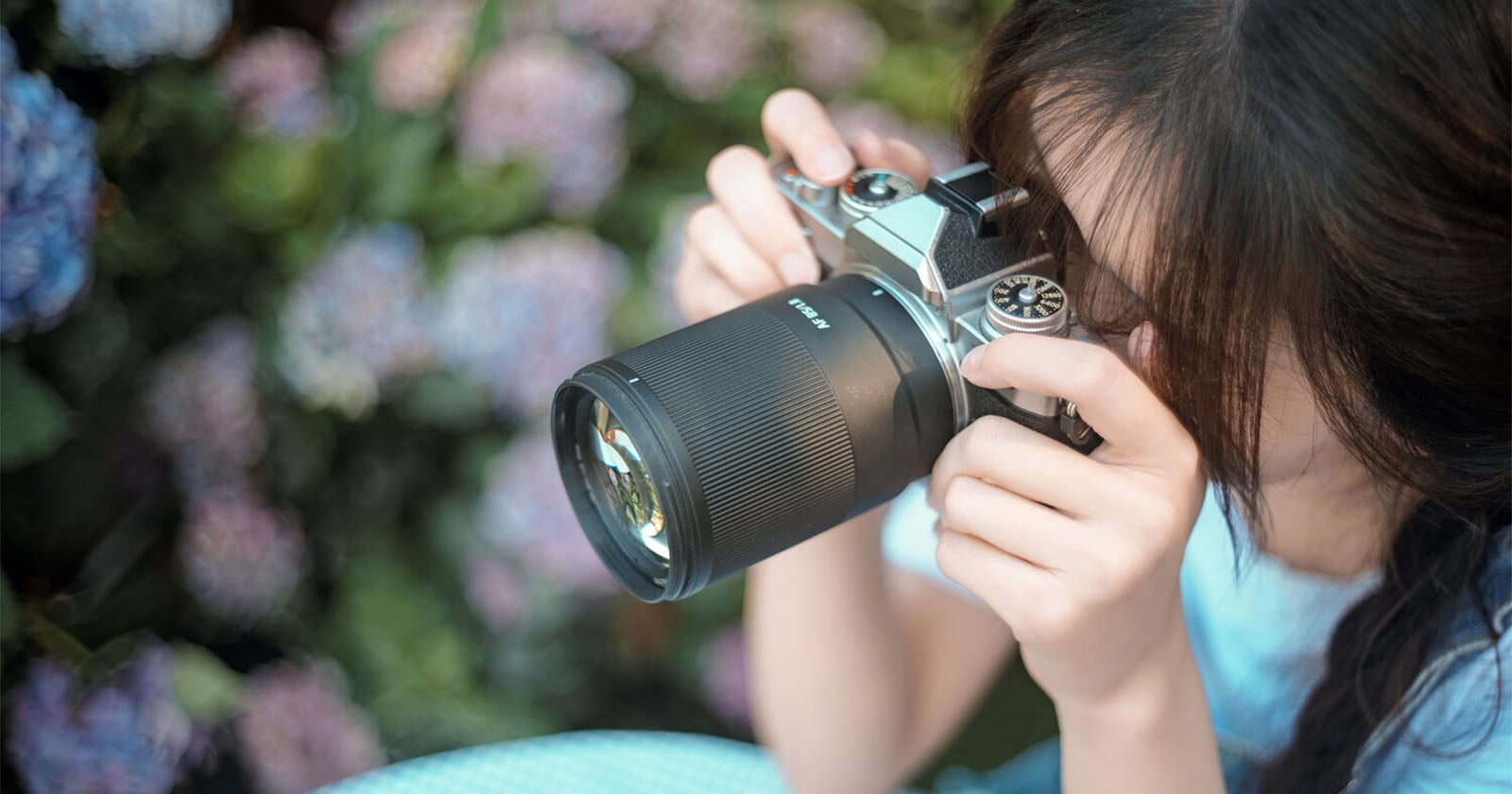 A person with dark hair holds a camera with a large lens up to their eye while standing in front of purple flowers.