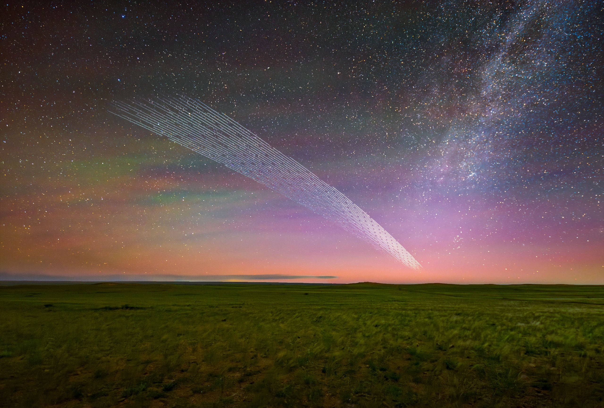 A long-exposure shot shows a train of satellite streaks arching across a night sky above a grassy landscape and the Milky Way.