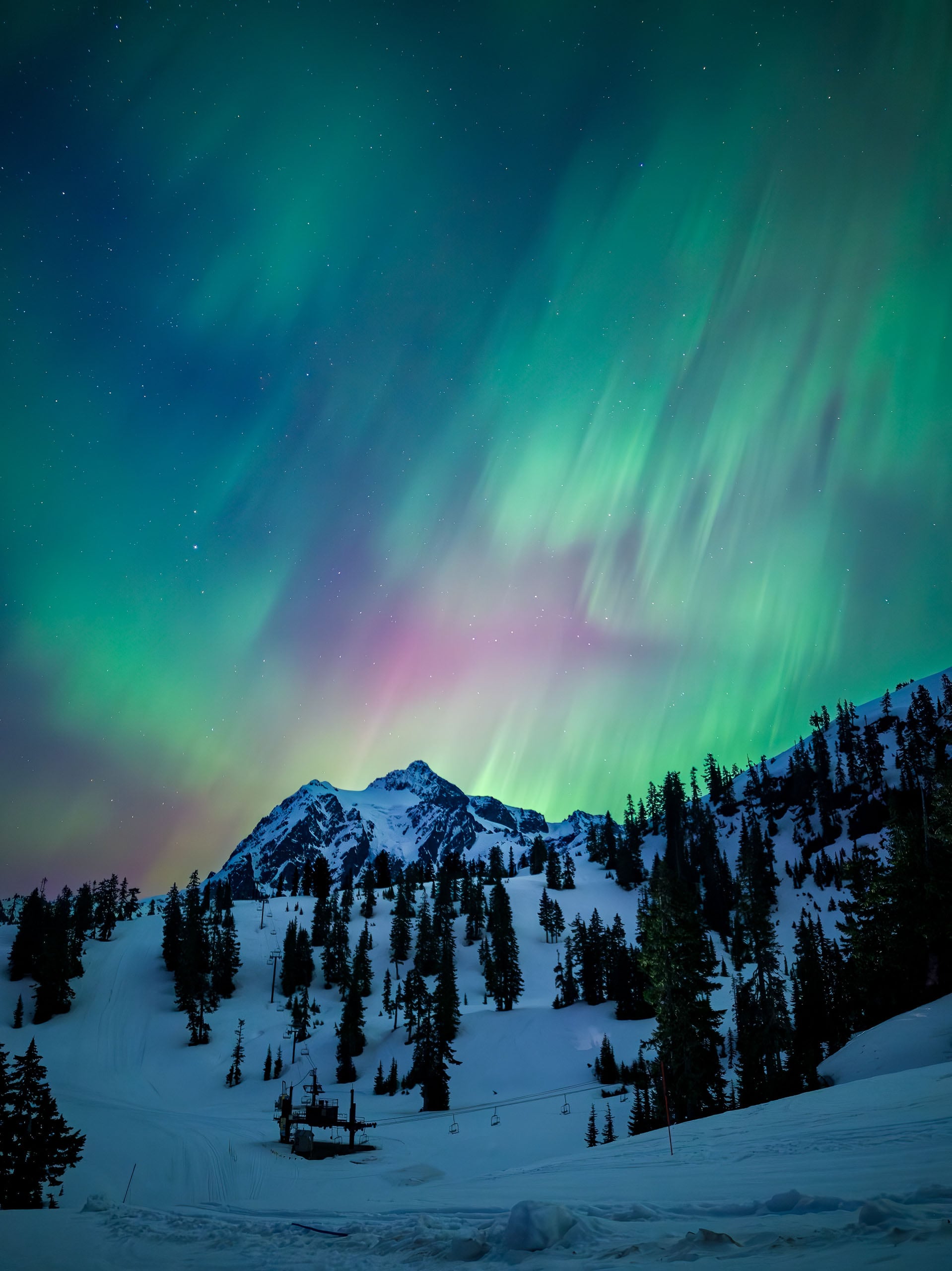 Vibrant green and purple aurora borealis illuminate the night sky above a snow-covered mountain slope with pine trees and a ski lift.