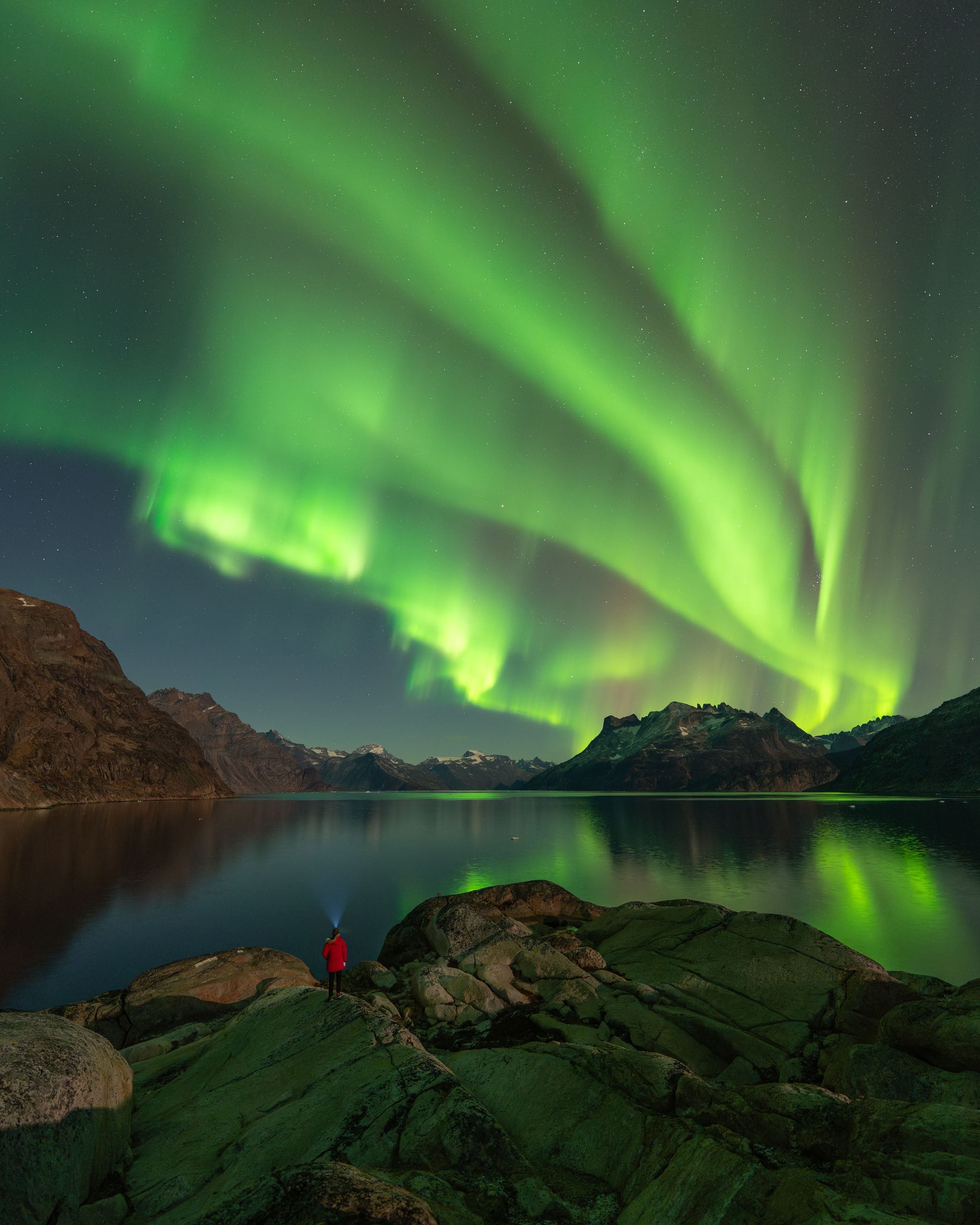 A person in a red jacket stands on rocky terrain at night, looking out over a calm lake toward mountains under a bright aurora.