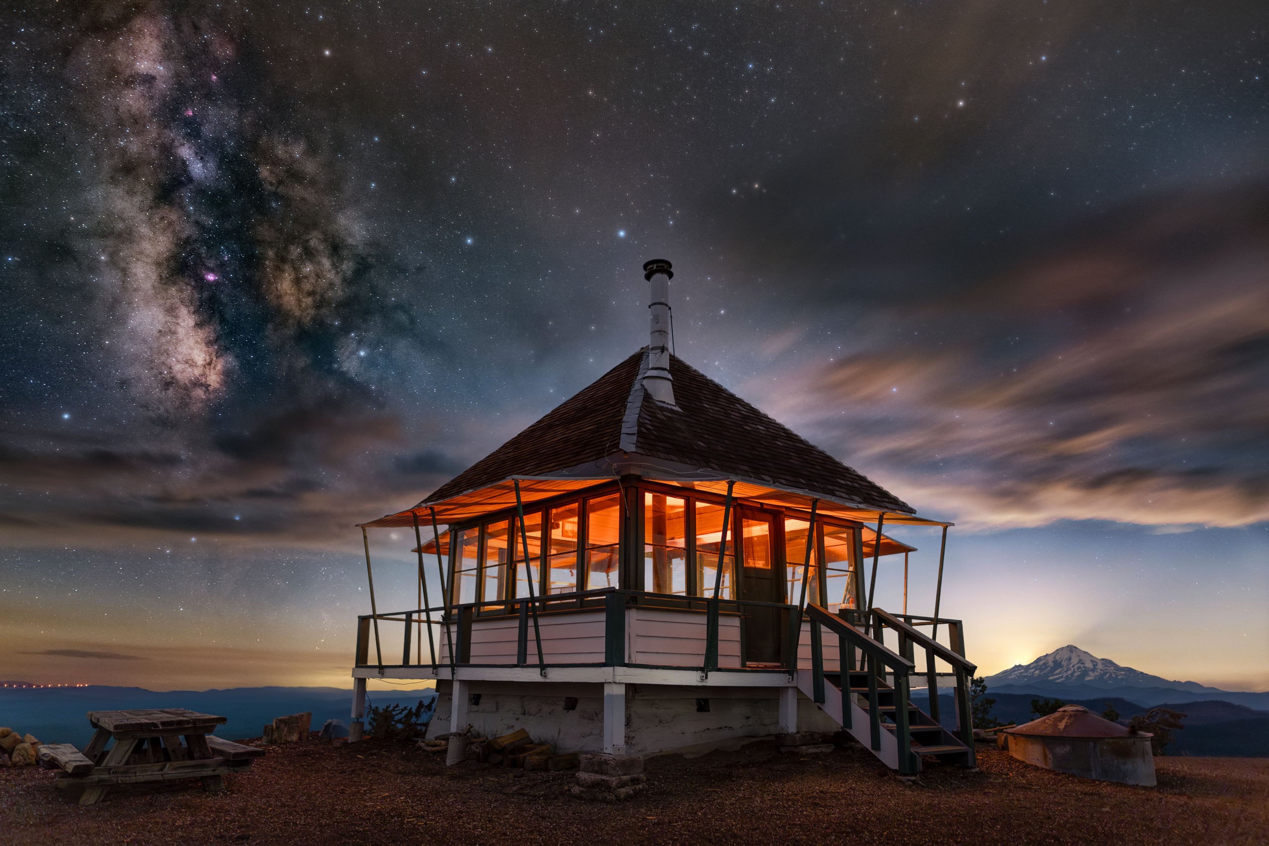 A wooden fire lookout cabin with glowing interior lights sits on a mountain peak under a starry night sky with the Milky Way.