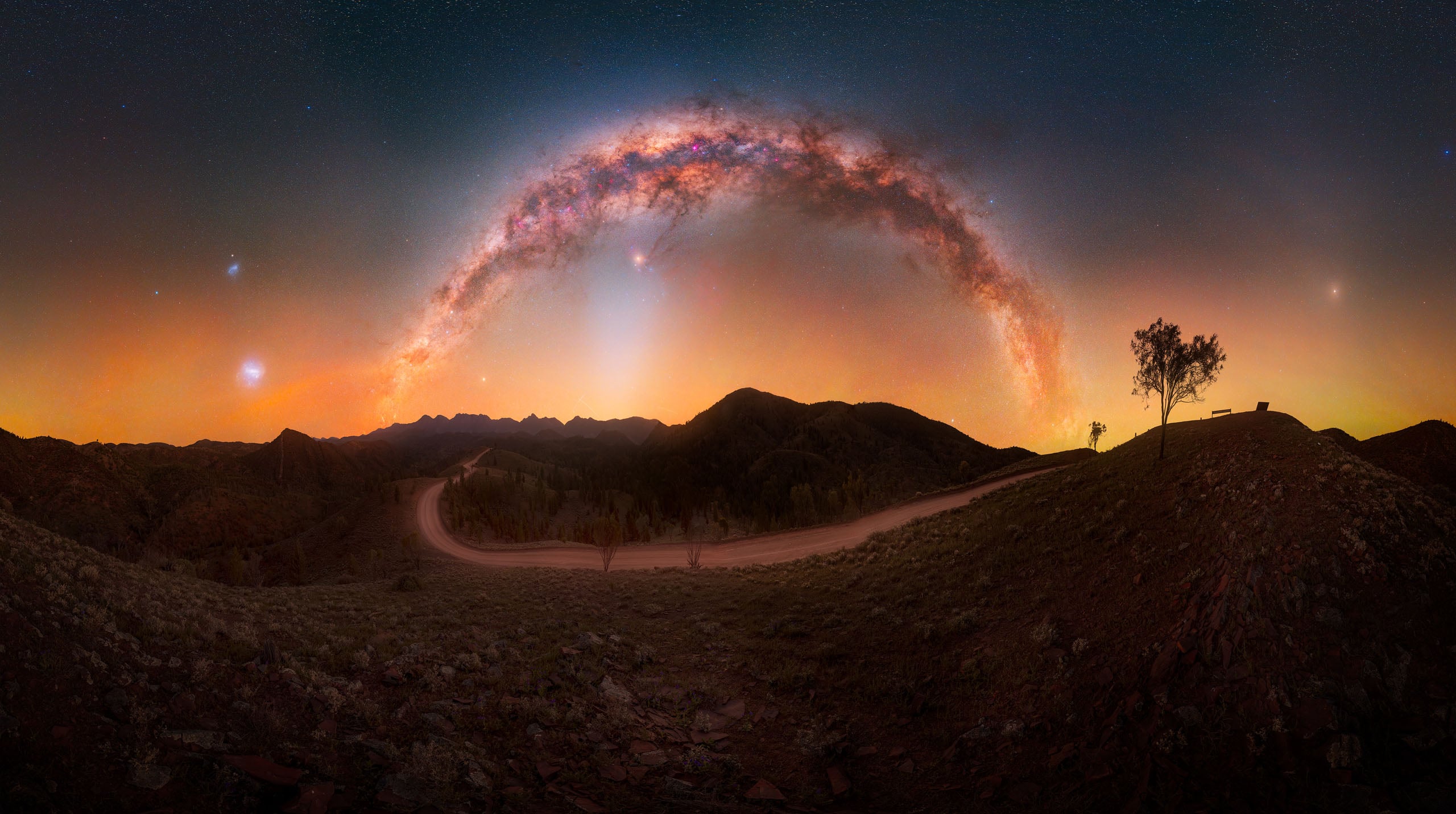 A panoramic view of the Milky Way arching over a dark, hilly landscape with a dirt road and a lone tree on a ridge.